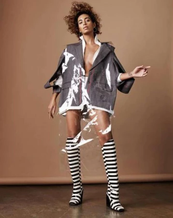 Fashion model posing in blazer, knee-high striped boots, and plastic skirt on a tan background in a studio, pointing to the right.