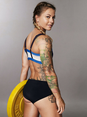 Athletic woman with tattoos in fitness wear poses in a studio holding a yellow weight plate, looking over her shoulder at the camera.