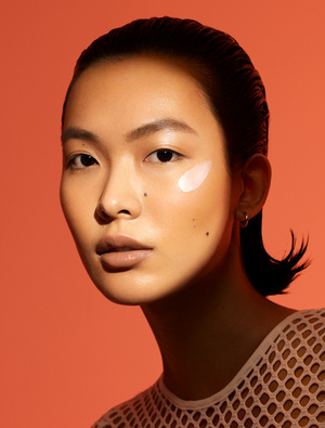 Close-up portrait of an East Asian woman with slicked-back hair and a cream smudge on her cheek against an orange background