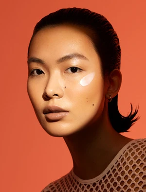 Close-up portrait of an East Asian woman with slicked-back hair and a cream smudge on her cheek against an orange background