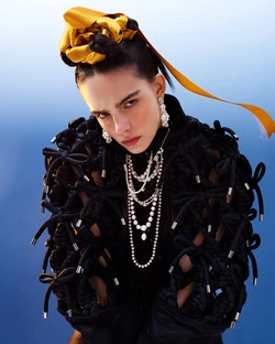 Portrait of a fashion model in a black jacket adorned with rope details, accessorized with a gold hair ornament, earrings, and multiple necklaces.