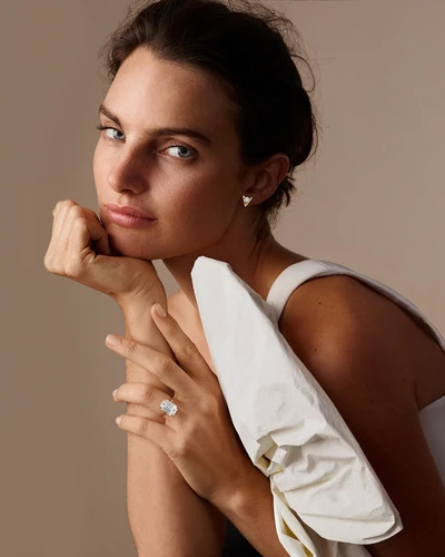 Close-up portrait of a young woman wearing a white top and a diamond ring, resting her face on her hand against a tan background.