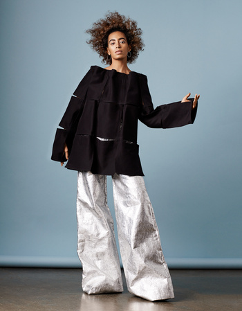 Young woman with curly hair poses in a black boxy top and metallic silver wide-legged pants against a light blue background.