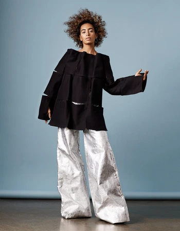 Young woman with curly hair poses in a black boxy top and metallic silver wide-legged pants against a light blue background.