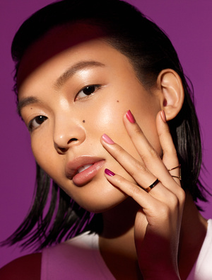 Close-up beauty shot of an Asian woman with two-tone pink manicure posing against a vibrant purple backdrop with dramatic lighting.