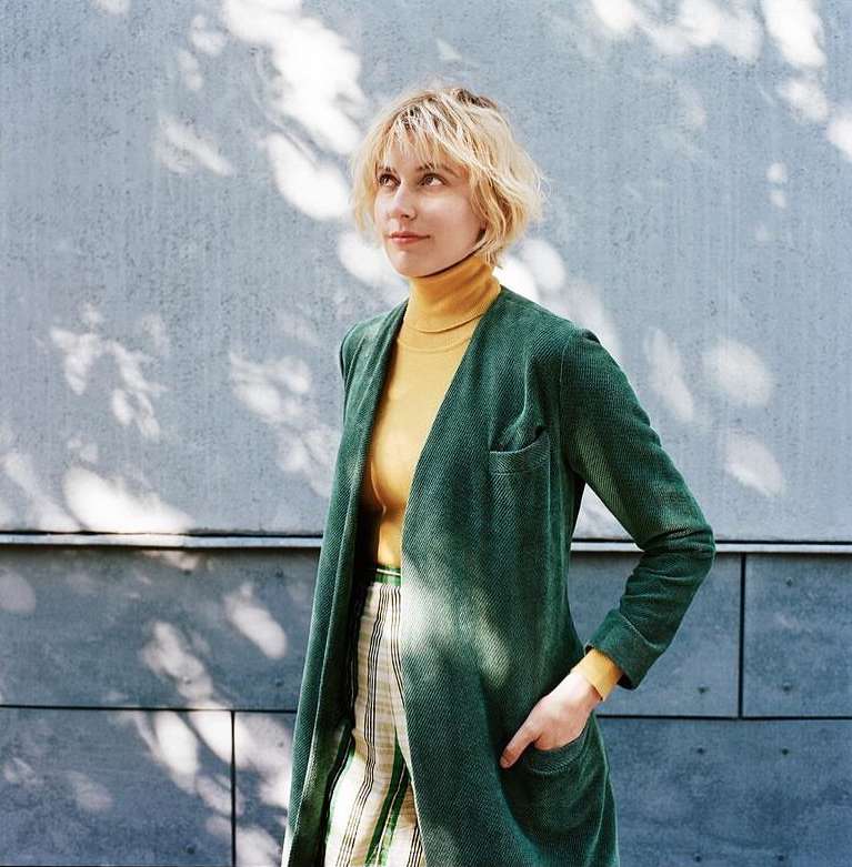 Adult female in green jacket with hand in pocket poses against concrete wall with shadows in an eye-level medium shot outdoors.