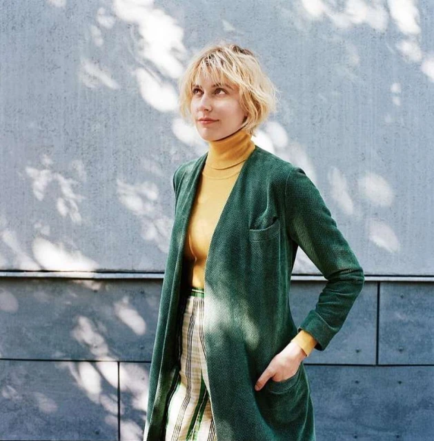 Adult female in green jacket with hand in pocket poses against concrete wall with shadows in an eye-level medium shot outdoors.