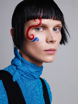 Close-up portrait of a woman with short black hair and a bold red and blue painted design on her face against a gray background.