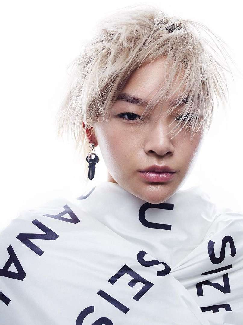 Close-up fashion portrait of a person with platinum blonde hair and a black key earring, wrapped in text-printed white fabric.