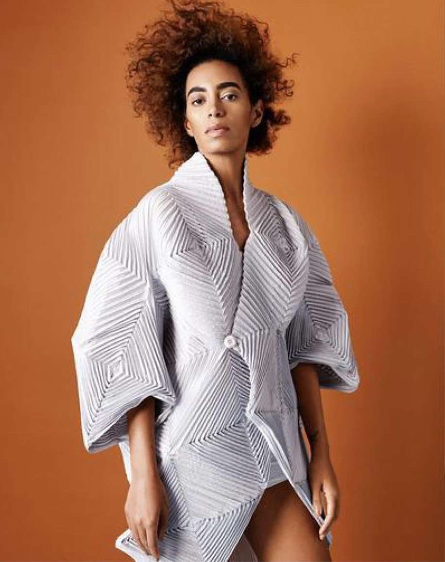 Fashionable adult female with curly hair, wearing an avant-garde white textured kimono-style jacket, posing against a warm orange backdrop.