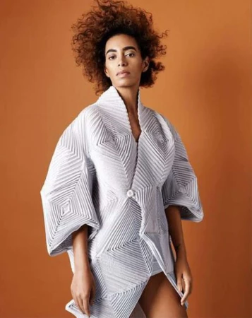 Fashionable adult female with curly hair, wearing an avant-garde white textured kimono-style jacket, posing against a warm orange backdrop.