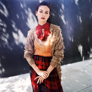 A woman in a red plaid skirt, tan shirt and cardigan stands outdoors against a dark wall with tree shadows in an eye-level medium shot.