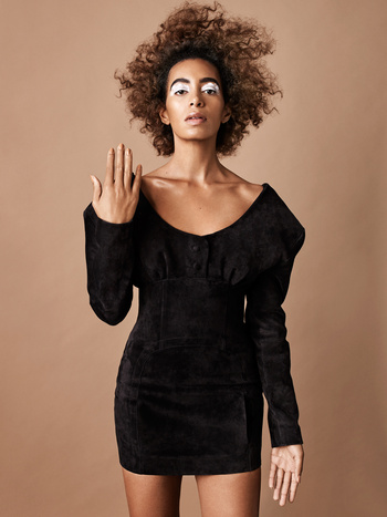 Portrait of a Black woman with voluminous curly hair wearing a black mini dress against a warm beige background.