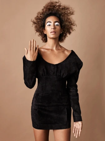 Portrait of a Black woman with voluminous curly hair wearing a black mini dress against a warm beige background.