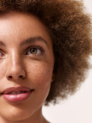 Close-up portrait of a woman with curly brown hair and freckled skin, her gaze directed slightly upwards in a beauty-style photograph.