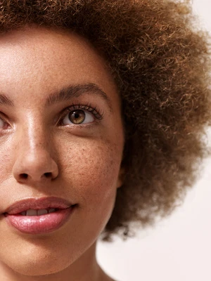 Close-up portrait of a woman with curly brown hair and freckled skin, her gaze directed slightly upwards in a beauty-style photograph.