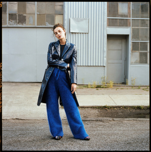Adult female poses on city street in black jacket and blue trousers, with neutral expression and natural lighting, looking at camera.