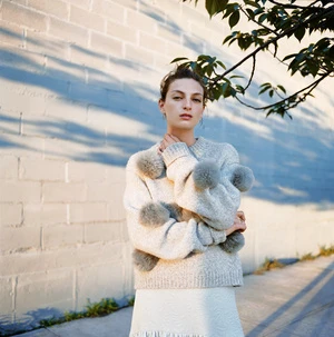 Model wearing a grey textured sweater with pom-pom details standing outside against a painted brick wall.