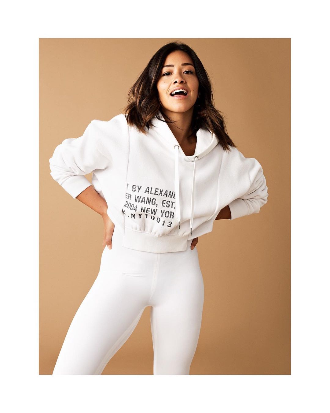Woman in white hoodie and leggings poses in front of a tan backdrop with an open mouth smile. Fashion shot, portrait framing.