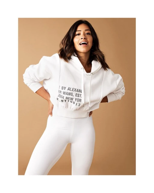 Woman in white hoodie and leggings poses in front of a tan backdrop with an open mouth smile. Fashion shot, portrait framing.