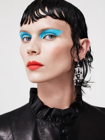 Close-up portrait of a model with wet-look dark hair, bright blue eye makeup, red lips, and geometric drop earrings.
