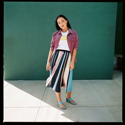 Woman wearing pink jacket and striped pleated skirt standing before a green wall