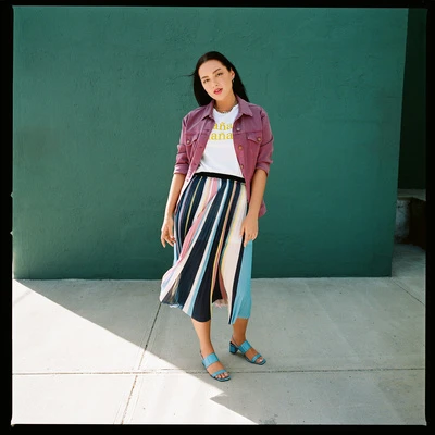 Woman wearing pink jacket and striped pleated skirt standing before a green wall