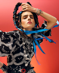 Portrait of a fashion model with intricate braids, wearing a patterned top against a vibrant red backdrop, showcasing a unique and bold style.