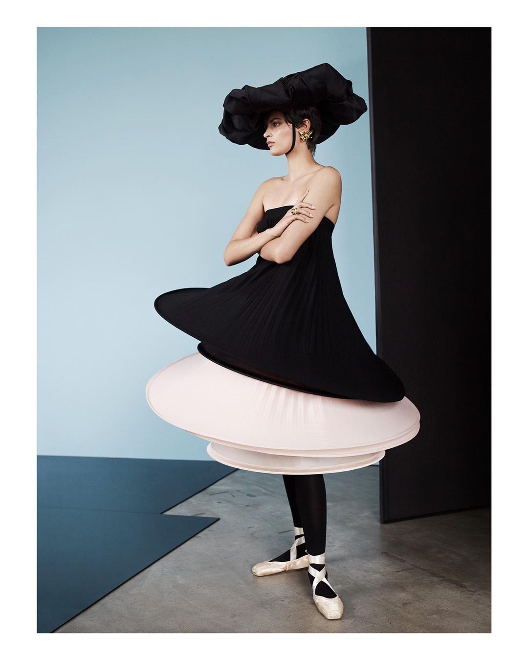 Young woman in couture black dress and oversized hat poses in a fashion studio with arms crossed, wearing ballet shoes, against blue wall.