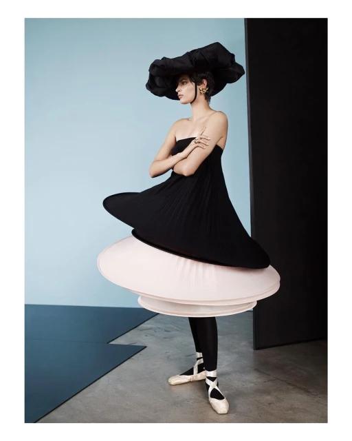 Young woman in couture black dress and oversized hat poses in a fashion studio with arms crossed, wearing ballet shoes, against blue wall.