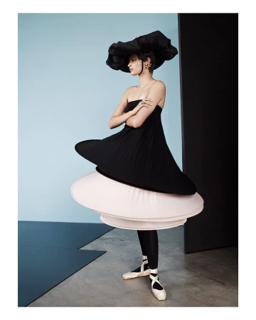 Young woman in couture black dress and oversized hat poses in a fashion studio with arms crossed, wearing ballet shoes, against blue wall.
