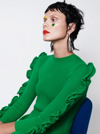 Model with dark wet-look hair, yellow eyebrow paint, and red lipstick wearing a green ruffled ribbed top against white.