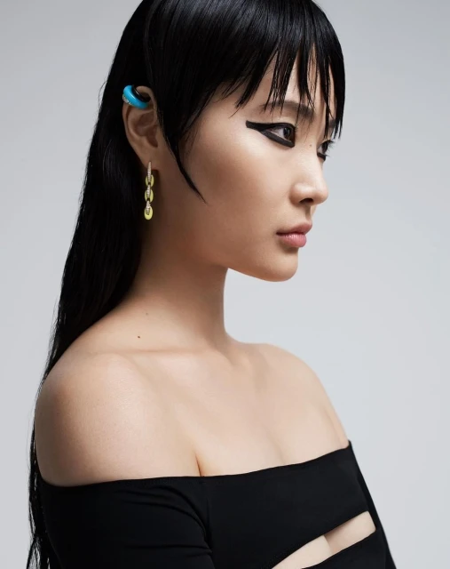 Glamour shot of an Asian woman with winged eyeliner and an ear cuff, posing against a gray background in a black, off-the-shoulder top.