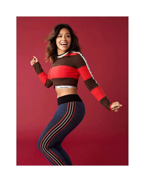 A young adult woman in athletic apparel poses dynamically against a red backdrop, exuding a playful and energetic mood in a studio setting.