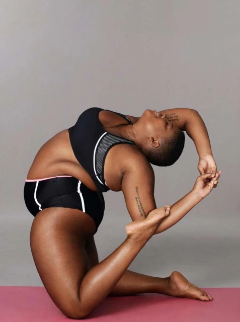An athletic woman with a shaved head is doing a yoga pose on a pink mat. She wears a black sports bra and black shorts in a studio.