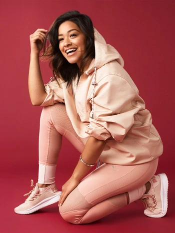 Gina Rodriguez poses kneeling indoors with a joyful expression, wearing athletic clothing against a solid red background.