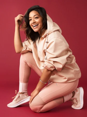 Gina Rodriguez poses kneeling indoors with a joyful expression, wearing athletic clothing against a solid red background.