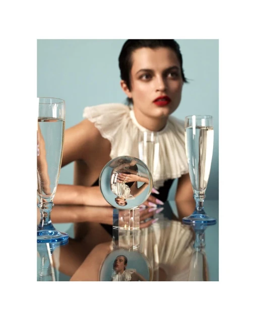 A fashionable female model with dark hair poses indoors with champagne glasses and a crystal ball on a reflective table against a blue backdrop.