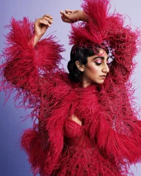 Woman in bright pink feathered jacket and matching bustier raising her arms against a purple gradient background