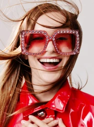 Close-up portrait of smiling woman wearing rhinestone-embellished red sunglasses and glossy red jacket with wind-blown hair against white background.