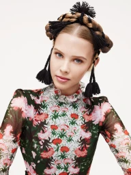 Portrait of a blonde woman wearing an elaborate braided crown hairstyle adorned with black tassels and a floral print high-neck dress.