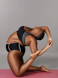 Plus size woman in a black sports bra and matching shorts performing a yoga backbend pose on a pink mat against a grey seamless backdrop.