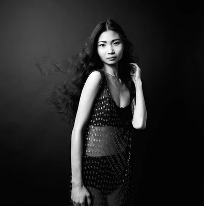 Monochrome portrait of an East Asian woman with long dark wavy hair wearing a patterned sheer dress against a dark background.