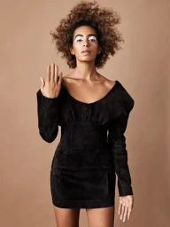 Portrait of a Black woman with voluminous curly hair wearing a black mini dress against a warm beige background.