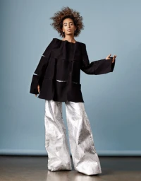 A woman with voluminous curly hair wearing a structured black top and shiny silver wide-leg trousers in a studio setting.