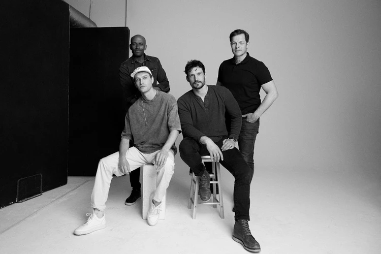Four men pose in a grayscale studio shot. One sits on a stool, another on a block, with two standing, showcasing a relaxed, casual group dynamic.