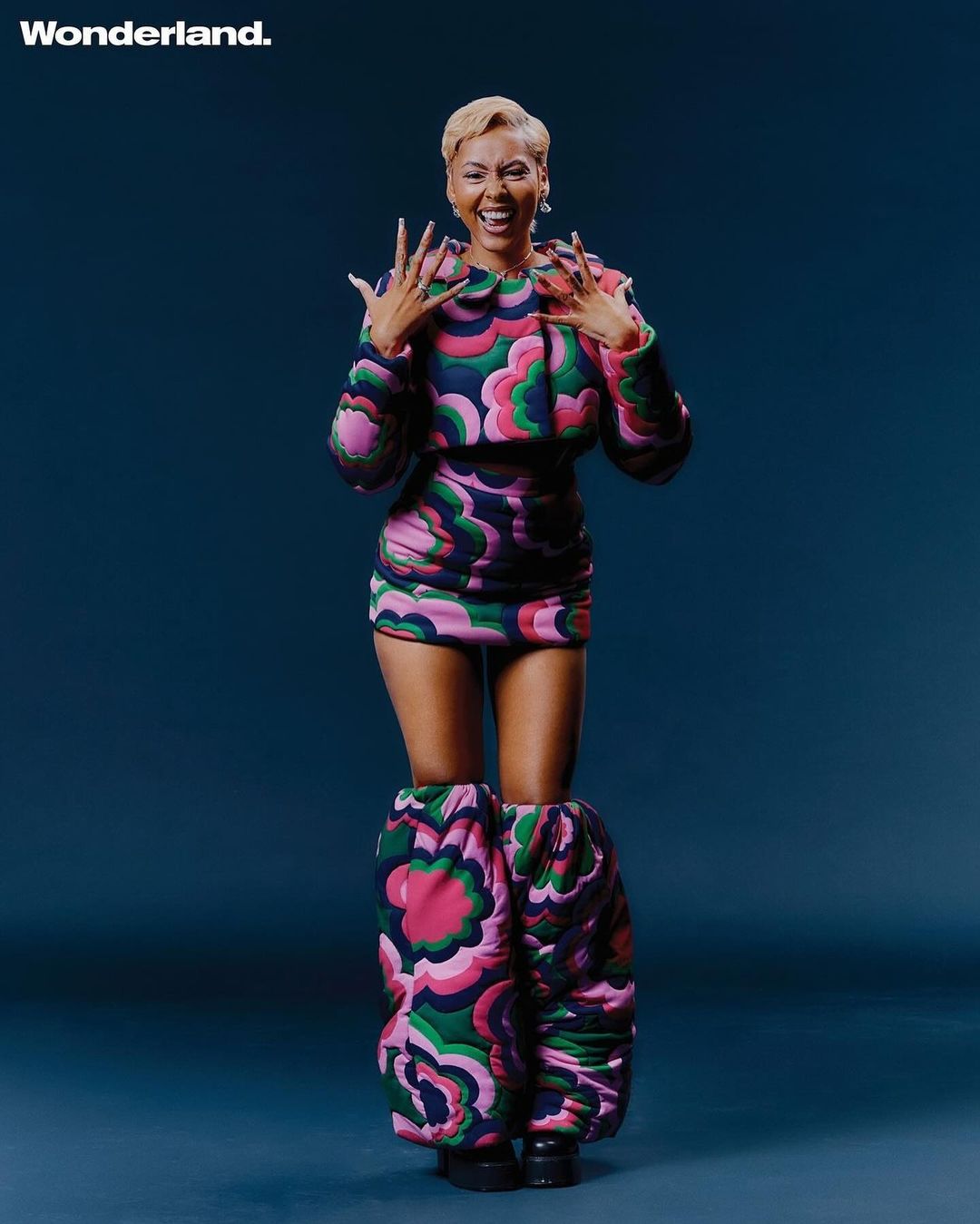 Woman with short blonde hair laughing in a matching floral patterned jacket, skirt, and oversized leg warmers.
