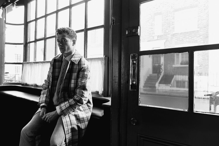 Man wearing a plaid coat seated inside near large windows by a street door, photographed by Stew Bryden.