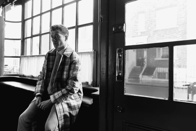 Man wearing a plaid coat seated inside near large windows by a street door, photographed by Stew Bryden.