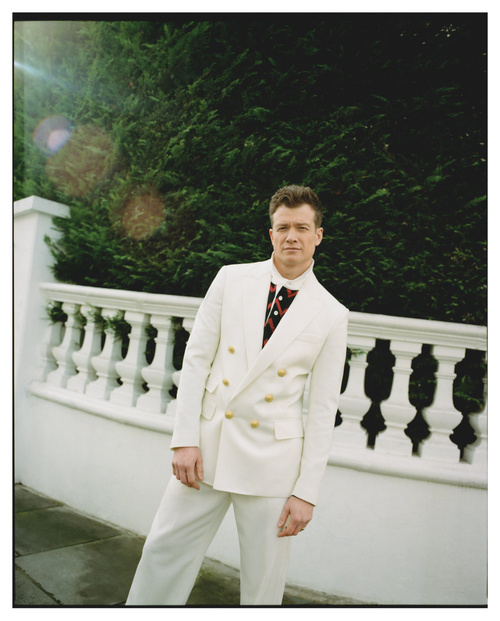 Man in an off-white double-breasted suit poses outdoors against a white balustrade with green foliage in the background.
